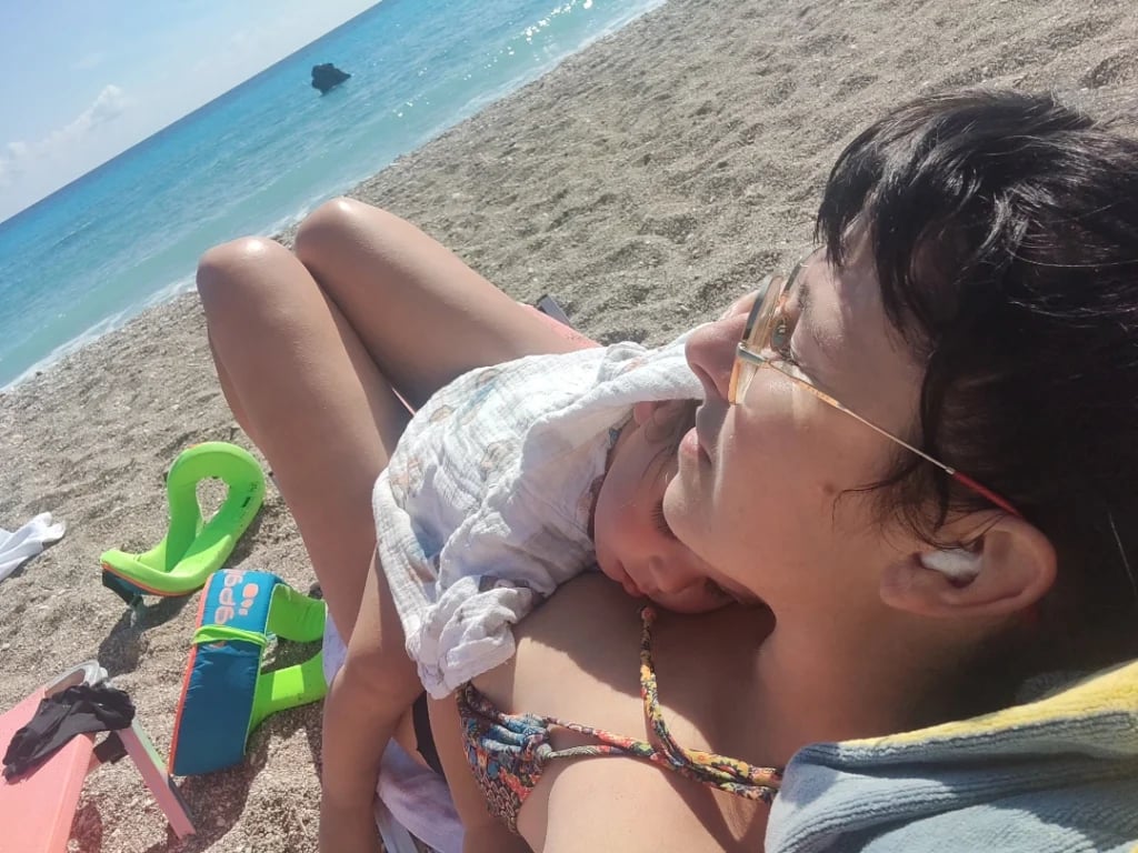 Majka se opušteno odmara na plaži pored mora dok joj beba spava na grudima